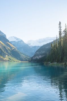 a lake surrounded by mountains and trees with blue water in the foreground on a sunny day