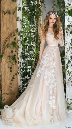 a woman wearing a wedding dress with long sleeves and flowers on the skirt is standing in front of an archway