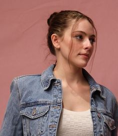 a woman wearing a jean jacket and white top standing in front of a pink wall