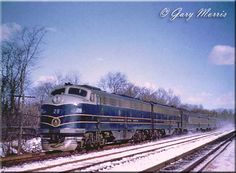 a blue and silver train traveling down tracks next to snow covered ground with trees in the background