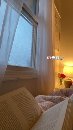 an open book sitting on top of a bed next to a window with white curtains