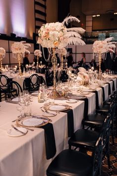 a table set up for a formal dinner with white flowers and feathers on it, along with black chairs