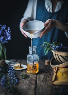 a person pours honey into a glass jar on a wooden table with blue flowers