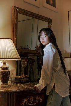 a woman standing next to a table with a lamp on top of it and a mirror behind her