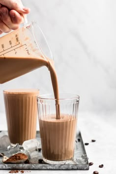 someone pouring chocolate into two glasses on top of a metal tray with coffee beans scattered around