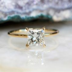 a close up view of a princess cut diamond ring on a white marble surface with a rock in the background