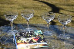 four wine glasses sitting on top of a glass table next to a book and some drinks