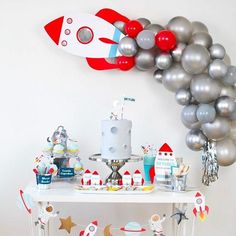 a birthday party with balloons, cake and other items on a white table in front of a rocket ship balloon wall