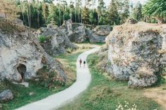 two people walking down a path in the middle of some large rock formations with trees on each side