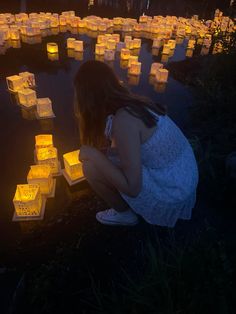 a woman kneeling down in front of some lit up paper lanterns on the water's edge