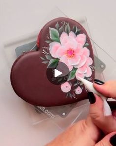 someone is painting a heart shaped box with flowers and leaves on the inside, while another hand holds a marker