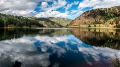 a lake surrounded by mountains under a blue sky with clouds in the middle and trees on both sides