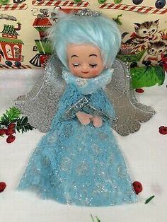 an old doll with blue hair is sitting on a white tablecloth next to christmas decorations