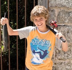 a young boy holding up a fish while standing in front of a fence with a fishing rod