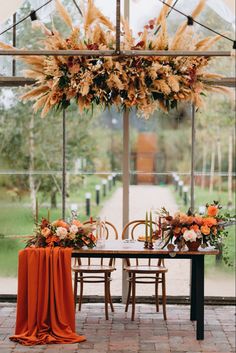 an orange table cloth draped over it with flowers and greenery in the center is surrounded by chairs