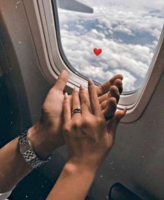 two hands reaching out an airplane window to touch the sky with a red heart on it