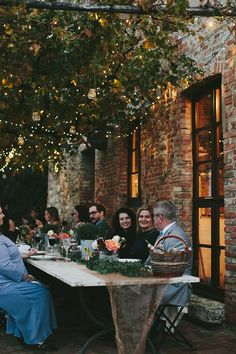 a group of people sitting around a wooden table with food and drinks on it in front of a brick building