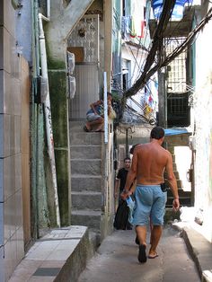 the man is walking down the narrow alleyway with his luggage in hand and no shirt on