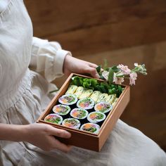 a woman is holding a wooden box with sushi in it and flowers on the side