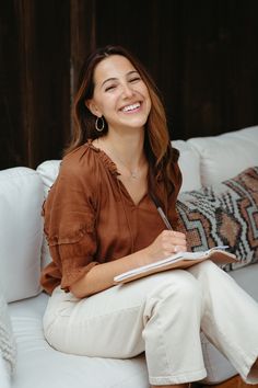 a woman sitting on a couch with a notebook in her lap and smiling at the camera