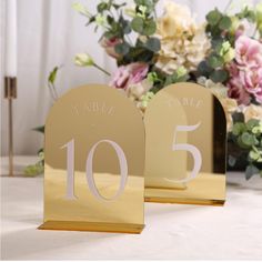 two gold table numbers are sitting on a table with flowers in the backgroud