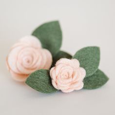 two pink flowers with green leaves on a white table cloth background, one is made out of felt and the other is made from wool