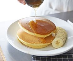 pancakes with syrup being poured onto them and sliced bananas on the plate next to them