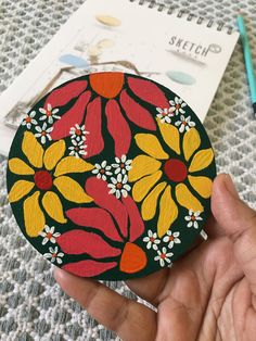 a hand holding up a painted flower on a piece of wood next to a notebook