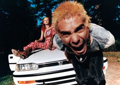 two women sitting on the hood of a white car and one man is sticking his tongue out