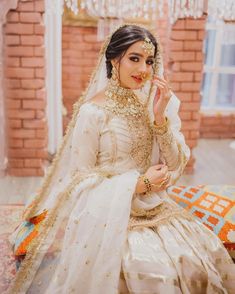 a woman in a white and gold bridal outfit sitting on the ground with her hand up to her face