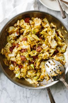 a pan filled with cabbage and bacon on top of a white marble counter next to utensils