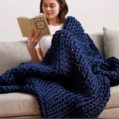 a woman is sitting on a couch holding a book and wearing a blue knitted blanket