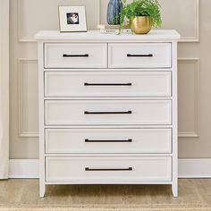 a white chest of drawers with plants and pictures on the top, against a beige wall