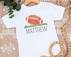 Elevate your little one's style game with our Toddler and Kid Football Shirt, a winning combination of sporty flair and personalized charm. Designed for comfort and individuality, this shirt features a prominent football graphic and can be customized with your child's name, creating a unique piece that's perfect for playtime, outings, and more. 🏈 Sporty Elegance: Our shirt showcases a meticulously designed football, capturing the excitement of the game in a stylish and kid-friendly way. The graphic adds a touch of athleticism to your child's wardrobe, sure to catch the eye of fellow sports enthusiasts. 👦👧 Perfect Fit for Toddlers and Kids: Available in a range of sizes to accommodate toddlers and older children alike, this shirt boasts a comfortable fit that allows for free movement dur Football Pro, Kids Football Shirts, Fall Sports, Style Sportif, Shirt Football, Personalized Football, Football Kids, Custom Football, Football T Shirt