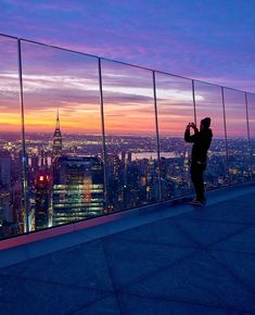 a man standing on top of a tall building next to a city skyline at sunset