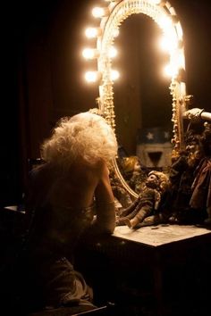 a woman sitting at a table in front of a mirror with light bulbs on it