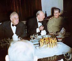 three men in suits sitting at a table with a cake and wine glasses on it