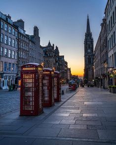 three red telephone booths sitting on the side of a street next to tall buildings at sunset