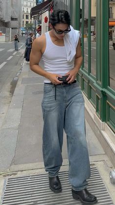 a man standing on the sidewalk looking at his cell phone while wearing jeans and a tank top