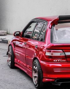 a red car parked on the street next to a white wall and cement flooring