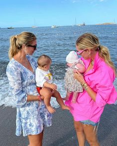 three women are standing on the beach and one is holding a baby while the other holds an infant