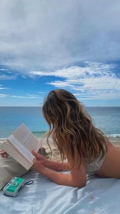 a woman laying on top of a bed reading a book next to the ocean and holding an mp3 player