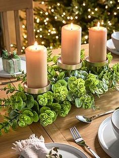 a table set for christmas with candles and greenery on the centerpiece, along with plates and utensils