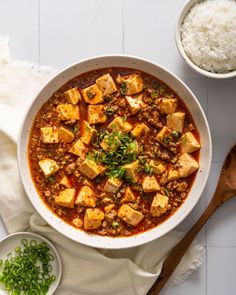 a white bowl filled with tofu and rice on top of a table next to a wooden spoon