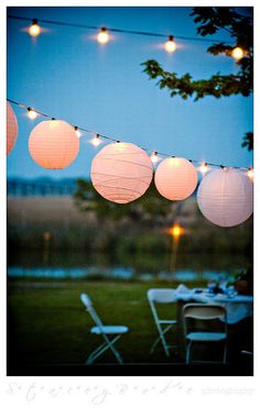 some paper lanterns are hanging from a string over a table with chairs and tables outside