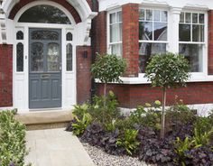 a house with red brick, white trim and green plants in the front garden area