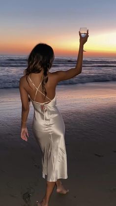 a woman is walking on the beach holding a wine glass in one hand and looking at the ocean