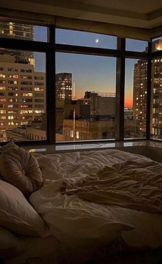 an unmade bed in front of a large window overlooking the city at night time