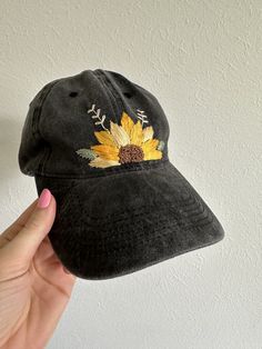 a black hat with a sunflower embroidered on the front and side, held up against a white wall