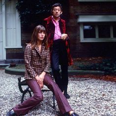 two people are sitting on a bench in front of a house and one person is standing